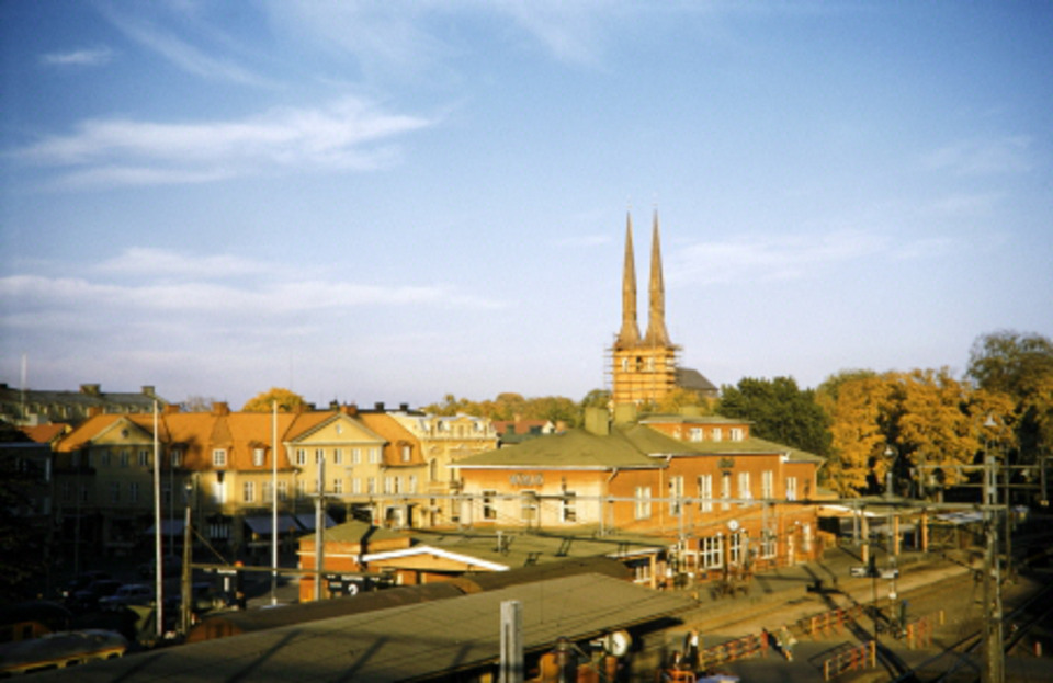 Vy över ett soligt Växjö en sensommardag från gångbron mot Växjö domkyrka. I förgrunden syns järnvägsstationen med stationshuset och bangården och till vänster skymtar en del av husen på Norra Järnvägsgatan.