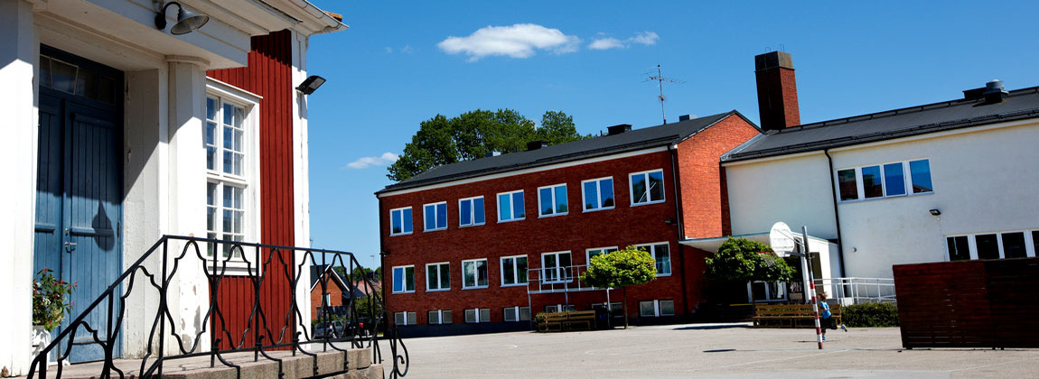 Röd trä byggnad med blå dörr, bredvid en röd flervåningsbyggnad i tegel. På skolgården springer en pojke i blå t-shirt